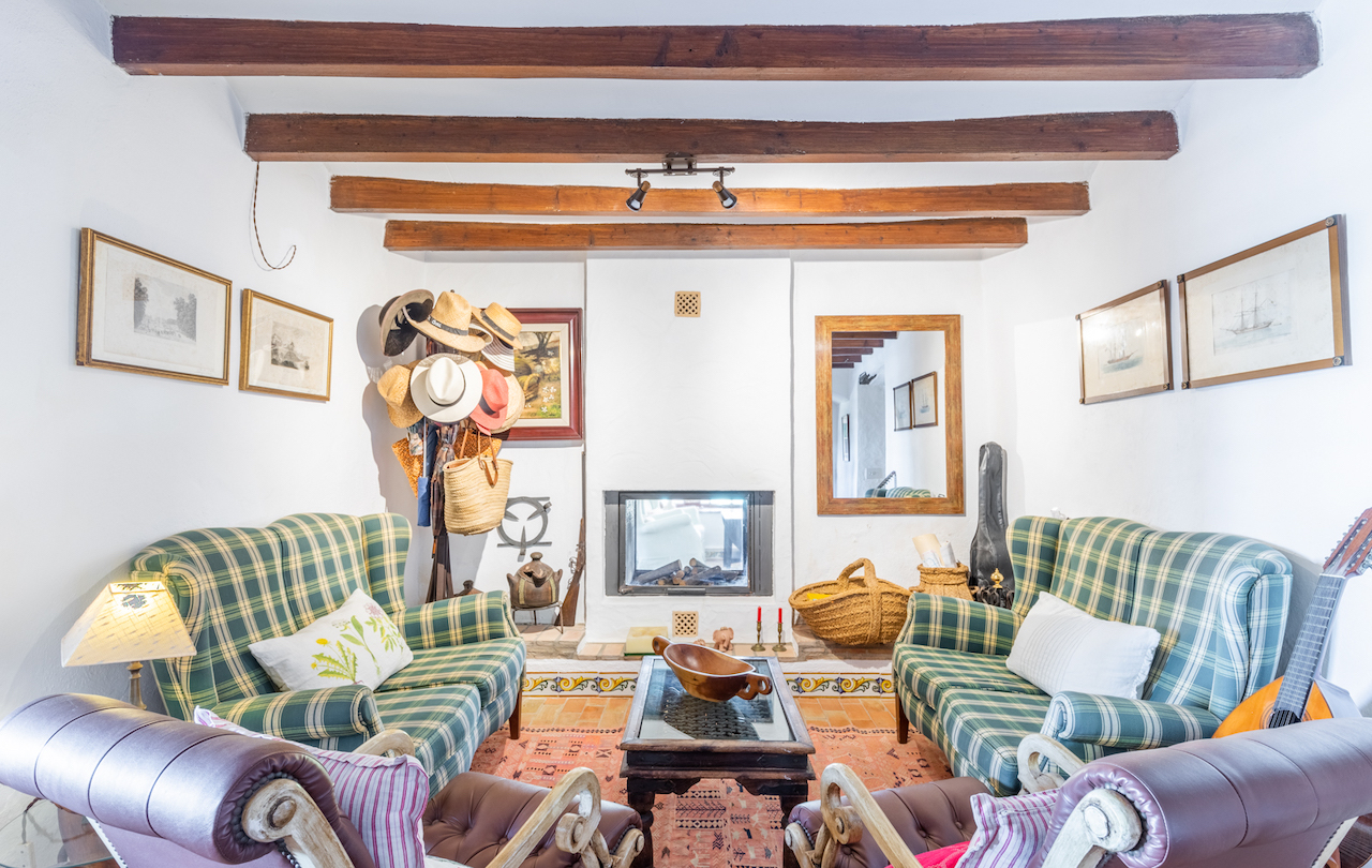 Rustic living area featuring exposed dark wood beams, a built-in glass fireplace, and terracotta flooring with decorative tiles. Furnished with plaid armchairs and traditional decor.