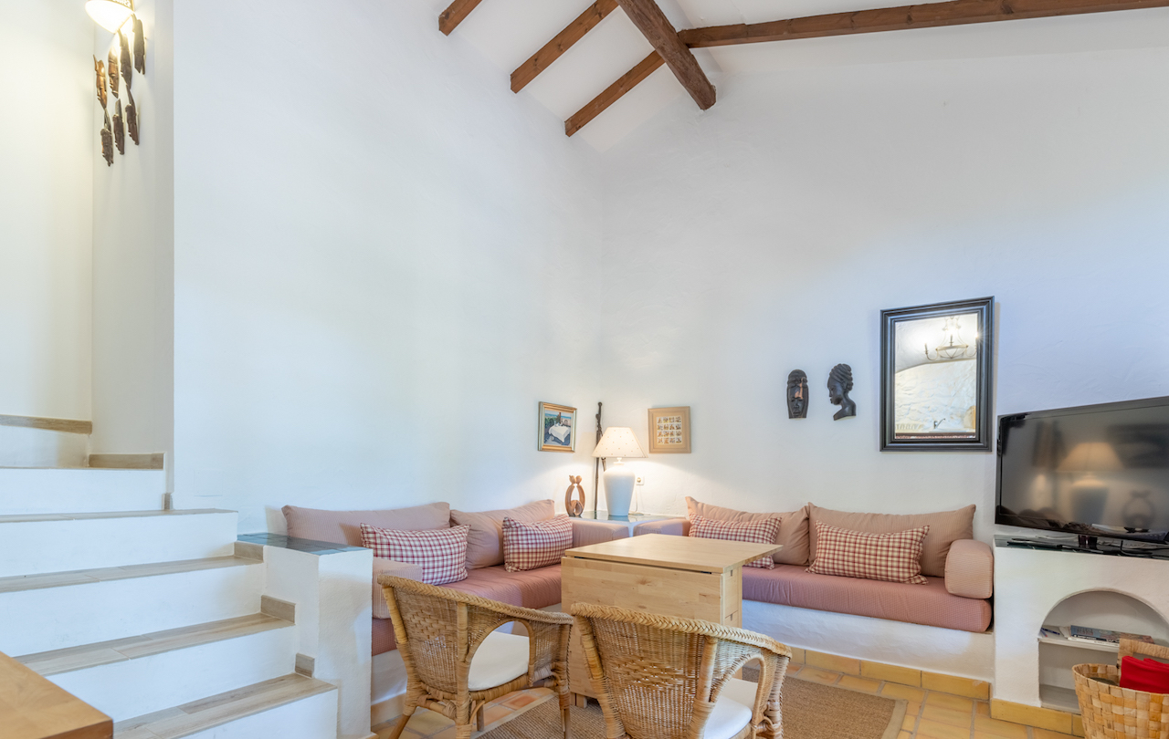 Rustic living area featuring high ceilings with exposed wooden beams, built-in seating with textile cushions, wicker armchairs, and traditional terracotta tile flooring.