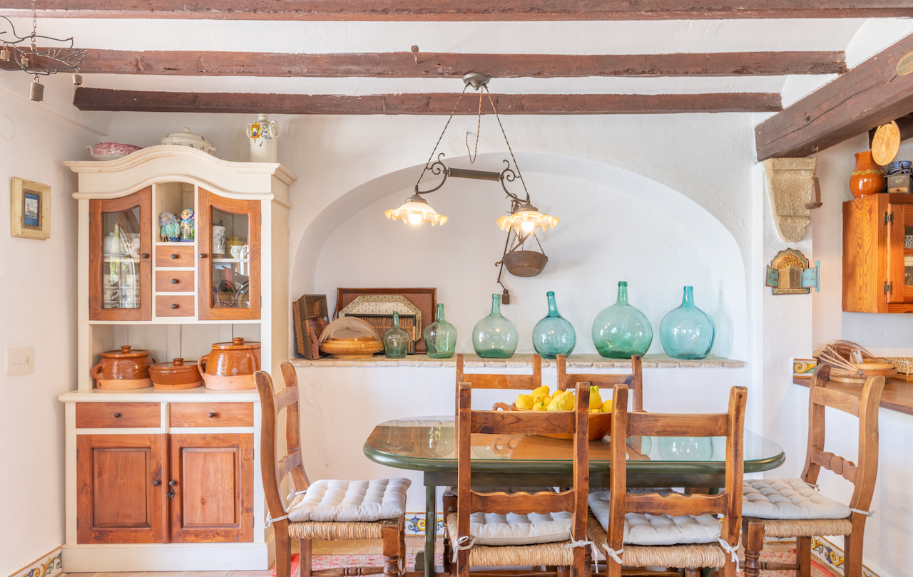 Rustic Valencian dining area featuring exposed wooden beams, a traditional hutch, and a built-in alcove displaying glass demijohns behind the dining set.