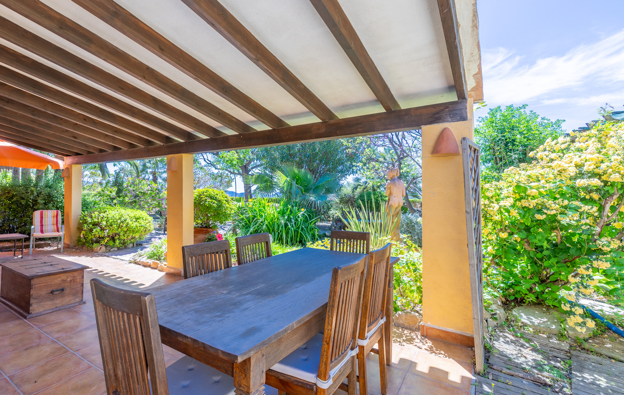Shaded terrace with traditional wooden beams and terracotta flooring, featuring a solid wood dining set overlooking a lush, manicured Mediterranean garden.