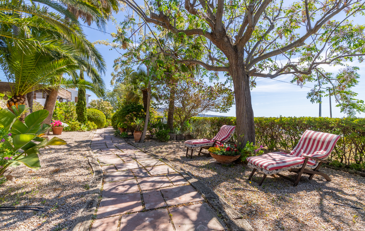Stone and timber pathway through a Mediterranean garden, featuring a shaded relaxation area with striped loungers beneath mature trees.