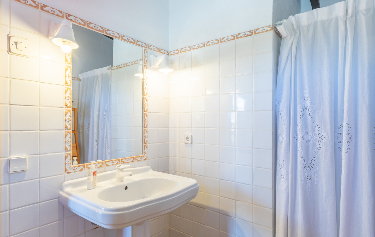 Traditional bathroom featuring a white pedestal sink, a mirror framed with decorative ceramic tiles, and classic wall sconces beside an embroidered curtain.