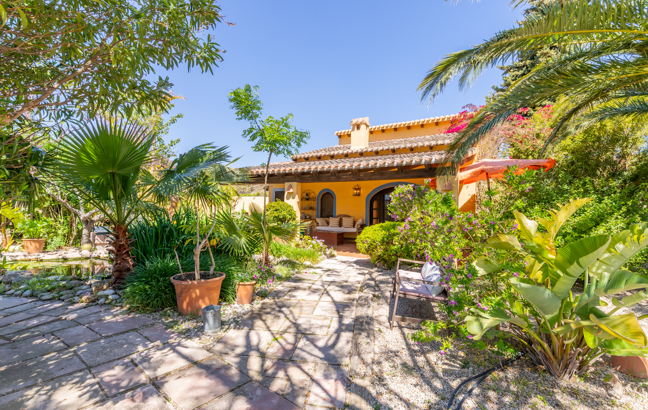 Traditional facade featuring a wooden-beamed porch, set within a lush Mediterranean garden with a stone path, palm trees, and a small pond.