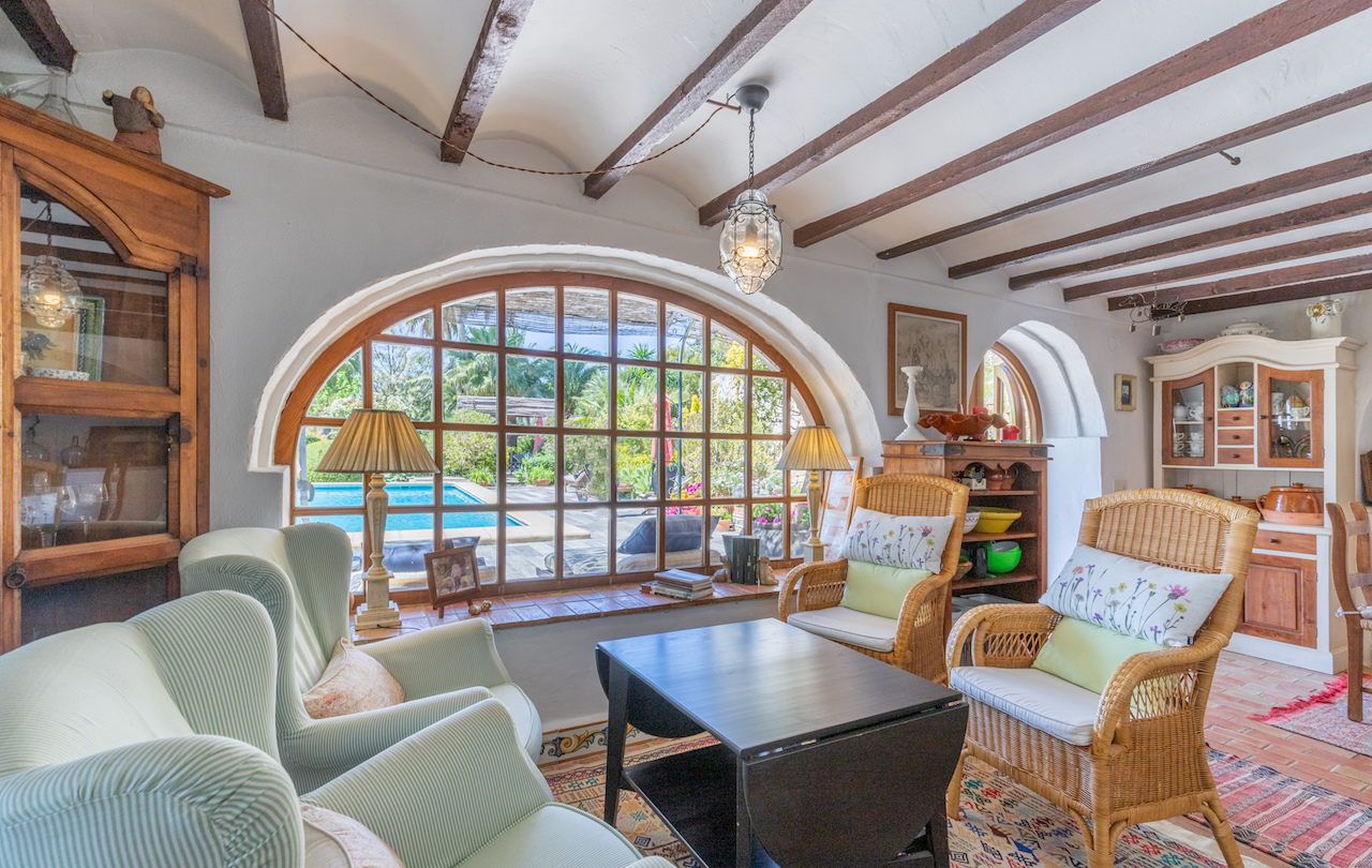 Traditional living area featuring exposed wooden beams, terracotta flooring, and a large arched window overlooking the swimming pool and lush gardens.