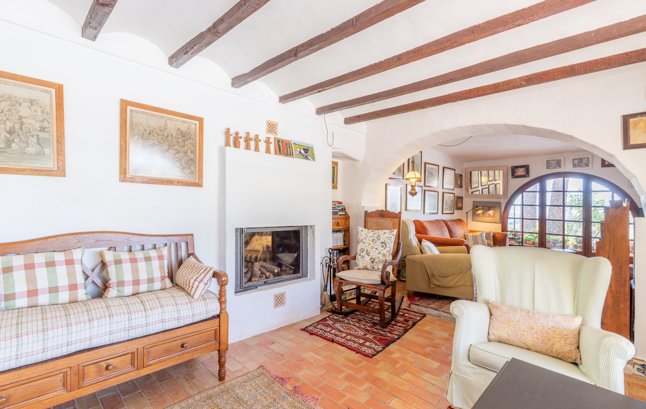 Traditional living room featuring exposed wooden beams, terracotta flooring, and a built-in fireplace. An archway leads to a bright seating area with a large arched window.