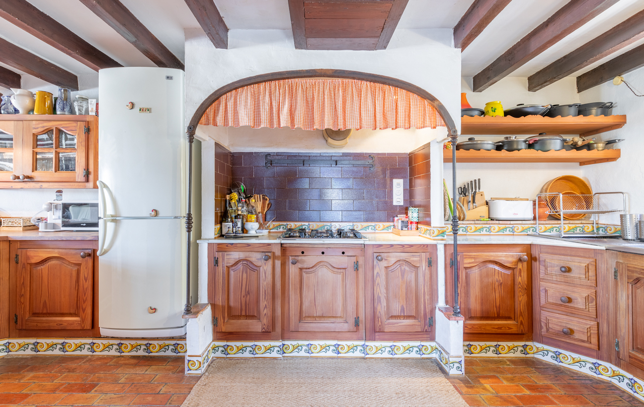 Traditional Valencian kitchen featuring exposed wooden ceiling beams, terracotta flooring, and a stove area framed by a decorative arch with artisanal tiling.