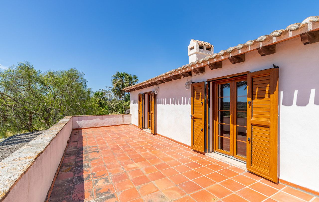 Upper terrace featuring terracotta tile flooring, exposed wooden eaves, and traditional wooden French doors with shutters against a white facade.