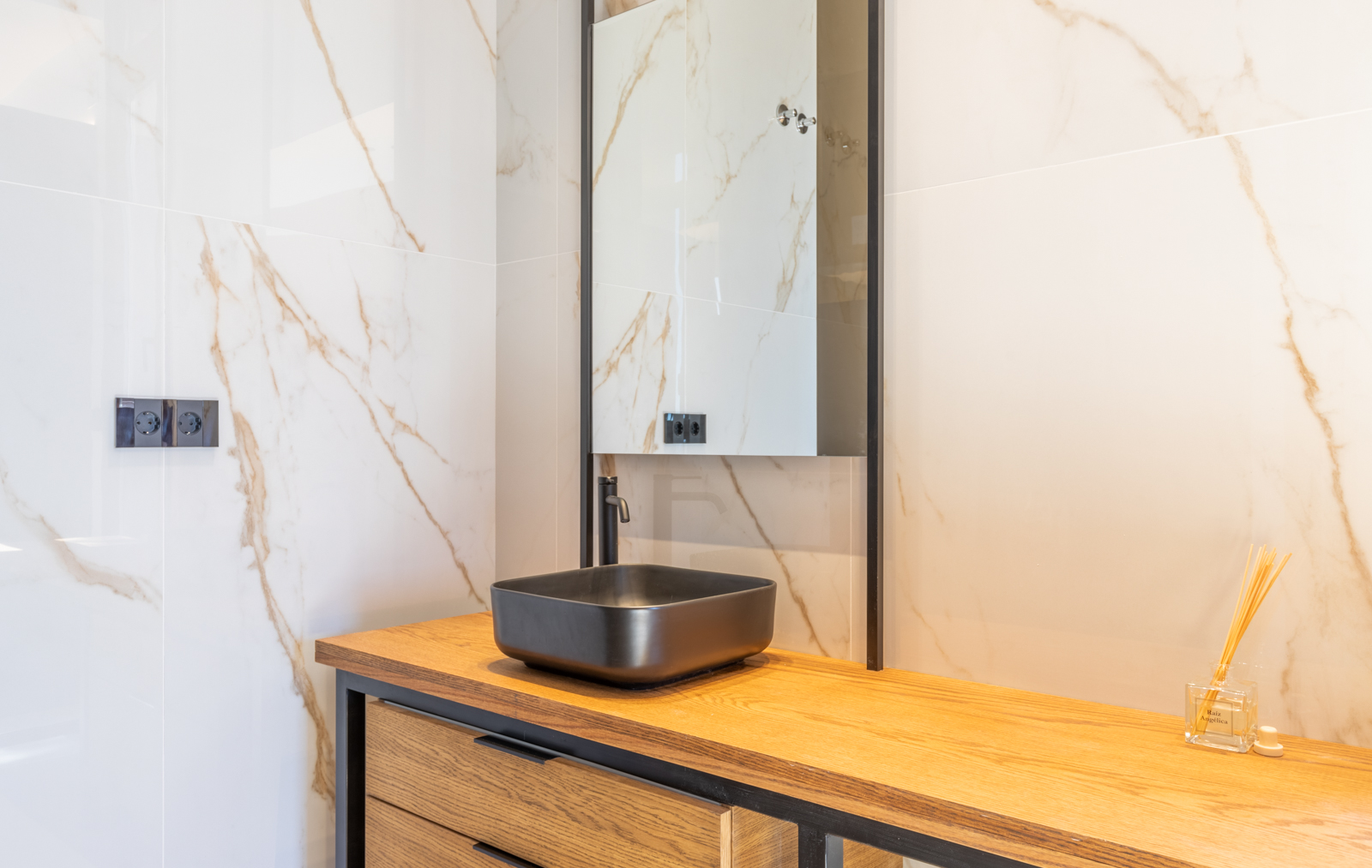 Bathroom detail with a matte black sink on a light wood countertop and a veined cream marble wall.