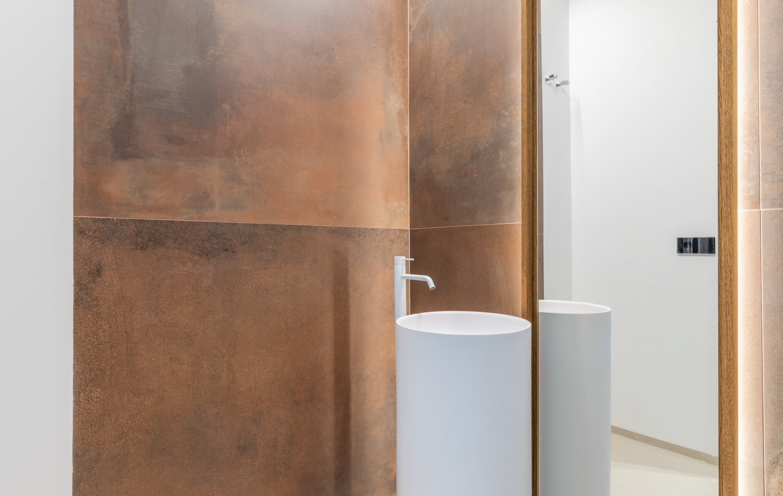 Bathroom detail with white cylindrical sink and rust-colored tiled wall with backlit mirror.