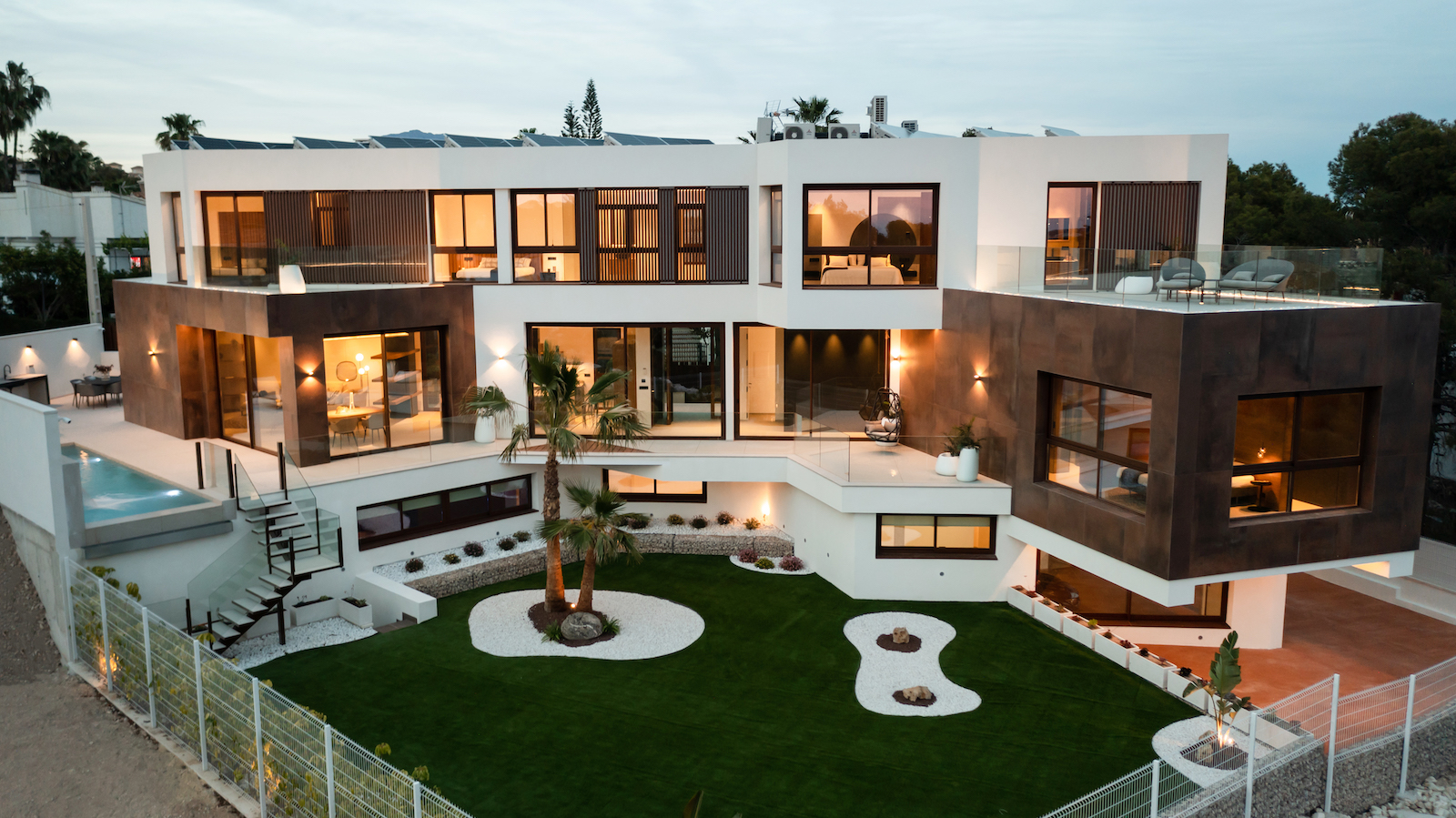Exterior view of the modern villa featuring a Mediterranean garden, pool, and illuminated terraces.