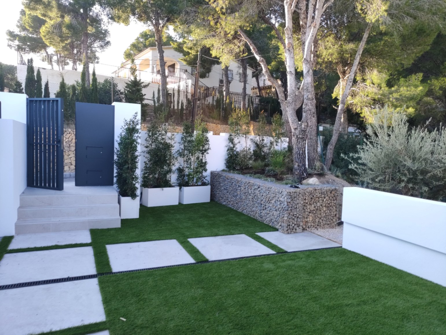 Exterior entrance featuring a modern black metal gate, stone steps, and a landscaped garden with artificial turf, concrete pavers, and a decorative gabion wall.