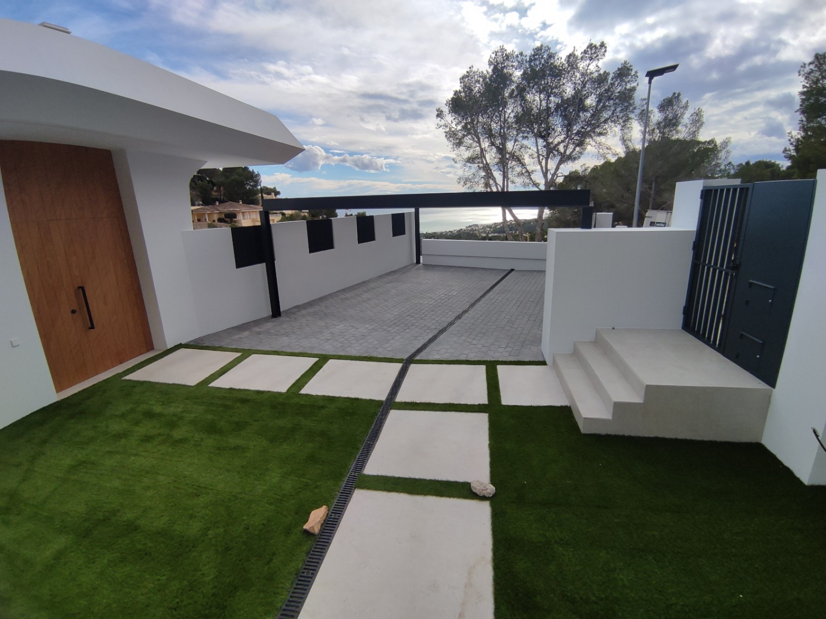 Exterior entrance featuring a solid wood door, paved driveway, and artificial turf with concrete pavers. The modern white structure overlooks the Mediterranean coast.