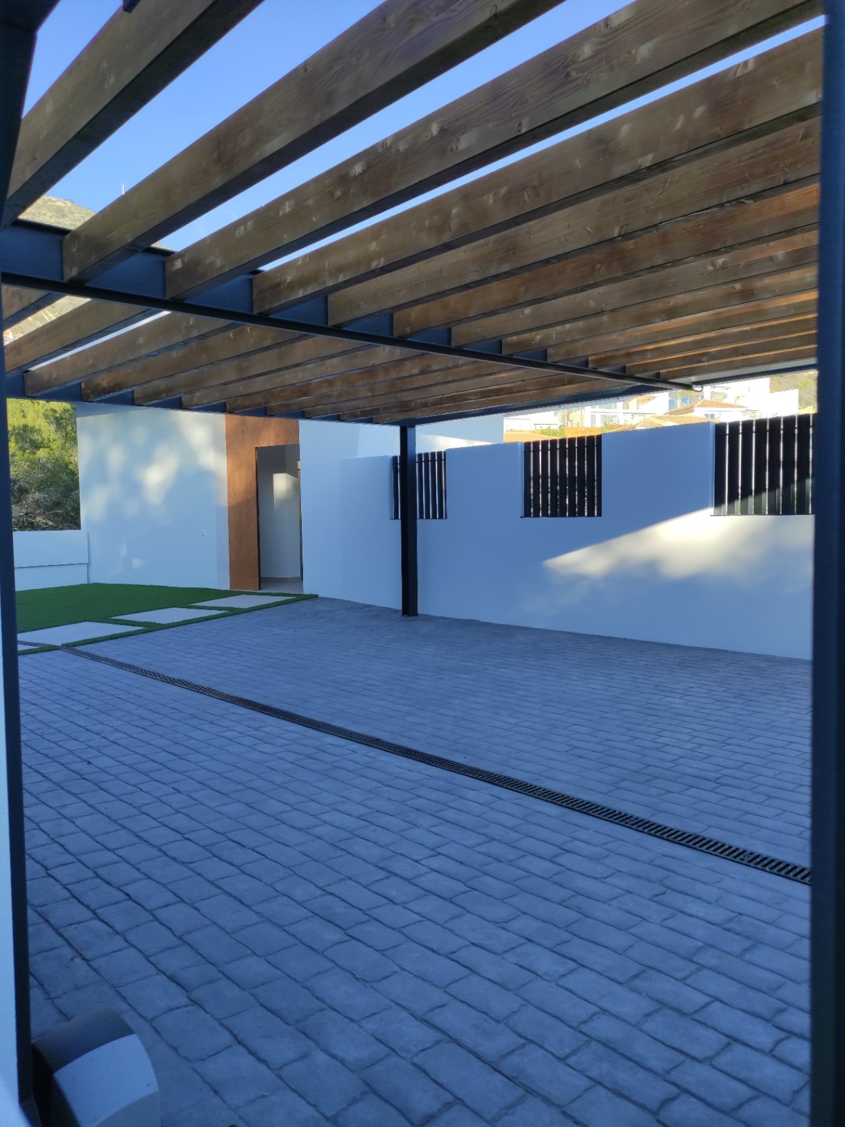 Exterior parking area featuring a wooden beam pergola, grey cobblestone paving, and a modern entrance with natural wood accents and white perimeter walls.