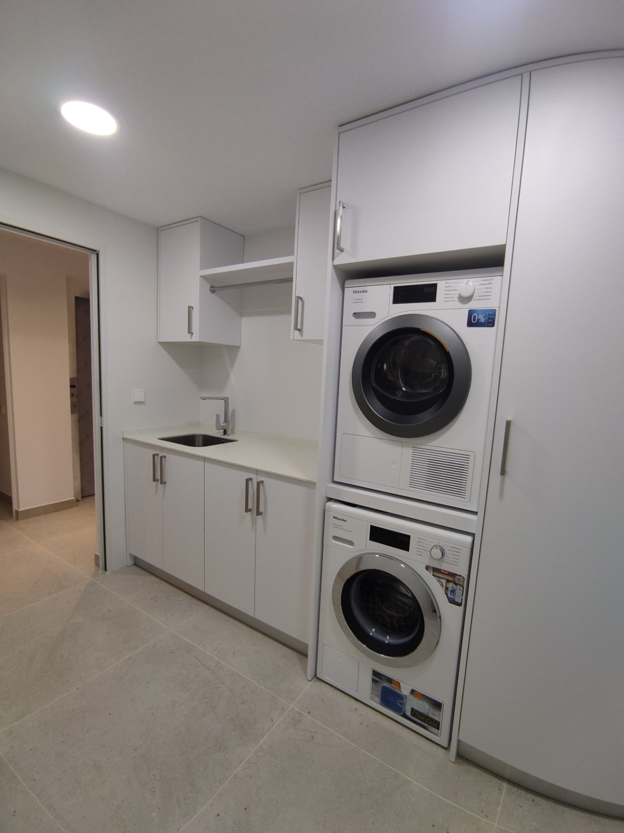 Functional laundry room featuring stacked Miele appliances, minimalist white cabinetry, an integrated sink, and neutral large-format floor tiles.