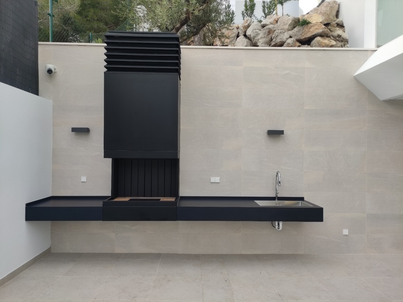 Minimalist outdoor kitchen featuring a matte black barbecue unit, integrated sink, and floating countertop set against a large-format stone wall.