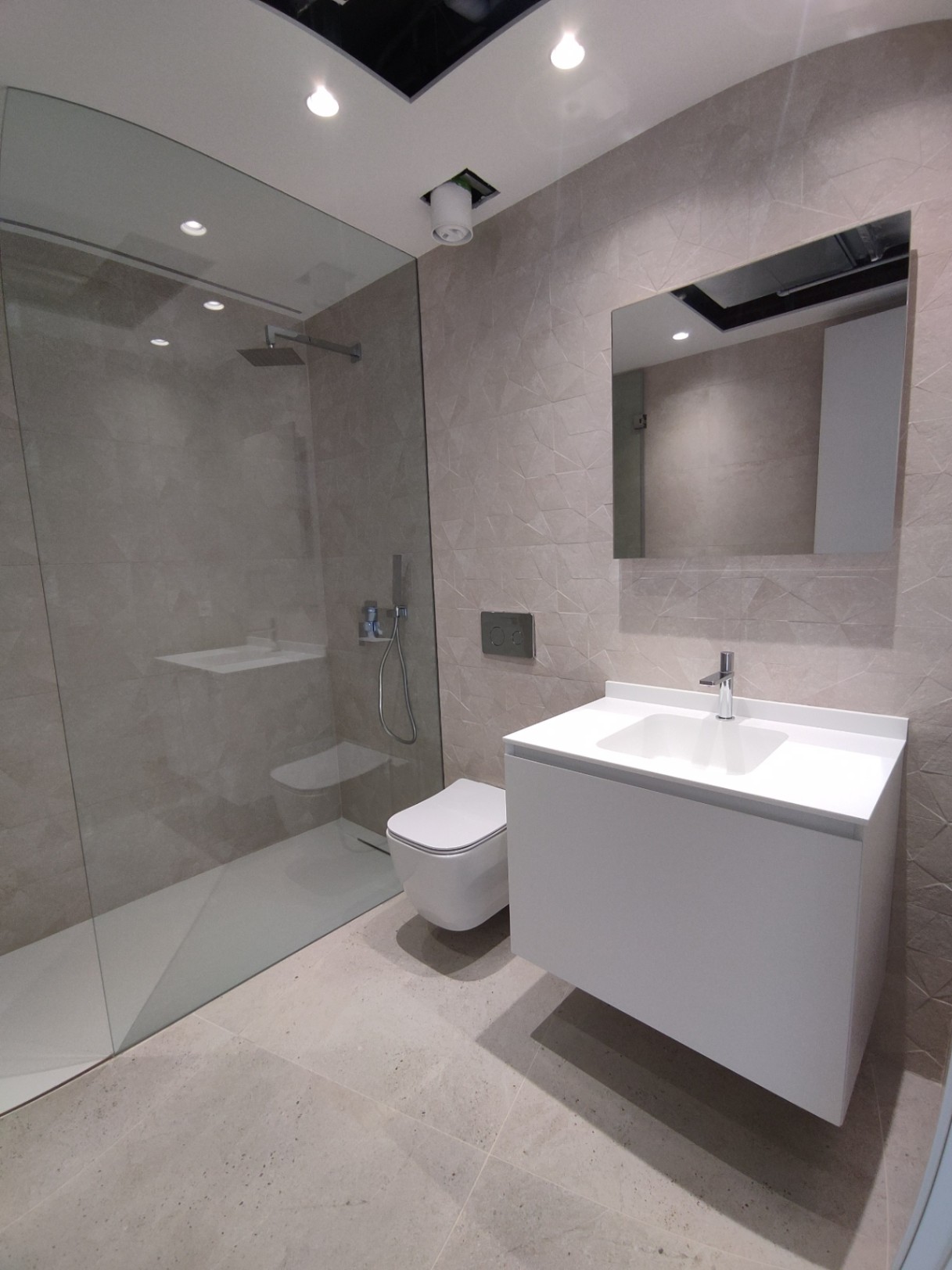 Modern bathroom featuring a walk-in glass shower, wall-hung toilet, and white floating vanity set against geometric textured wall tiles.