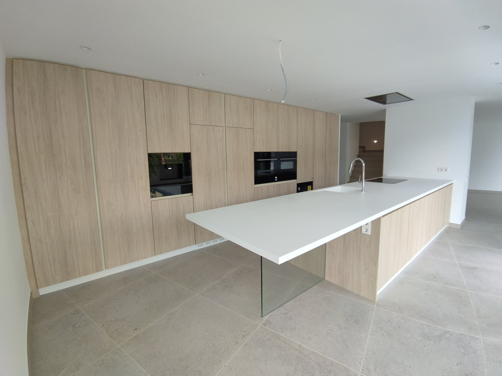 Modern kitchen featuring wood-finish cabinetry, integrated appliances, and a white island with a cantilevered glass support and porcelain flooring.