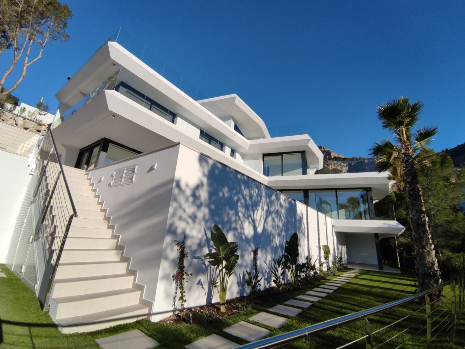 Multi-level contemporary exterior featuring white geometric architecture, glass railings, and a landscaped garden with a stone path and palm trees.