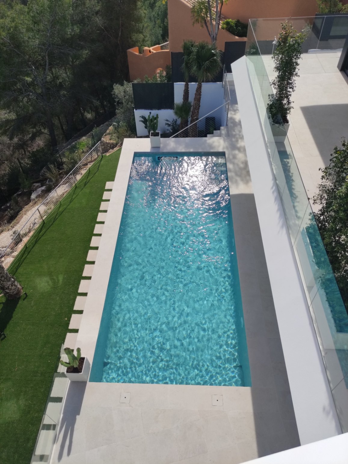 Rectangular swimming pool with turquoise water, featuring a light stone deck, integrated stepping stones on artificial turf, and modern glass balustrades.