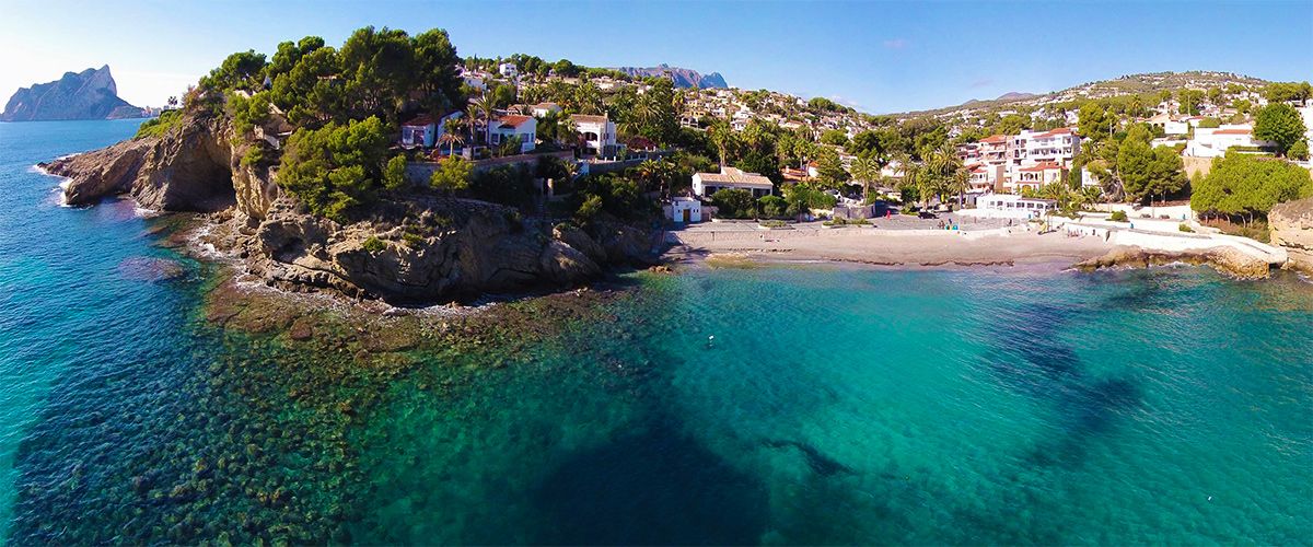 Aerial view of La Fustera beach featuring turquoise waters, rocky cliffs, and the Peñón de Ifach in the distance, set against a backdrop of pine trees and coastal villas.