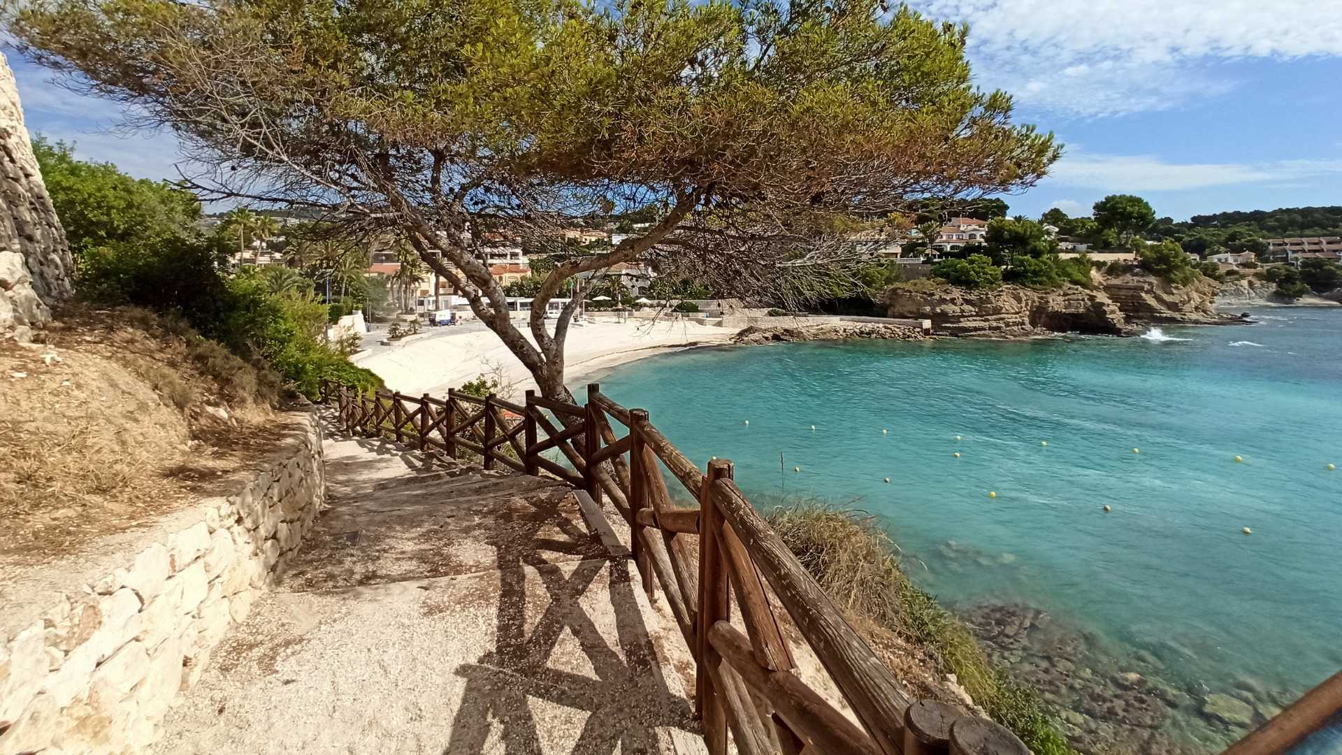 Coastal path featuring a wooden railing and natural stone walls, providing direct access to the sandy La Fustera beach and turquoise Mediterranean waters.