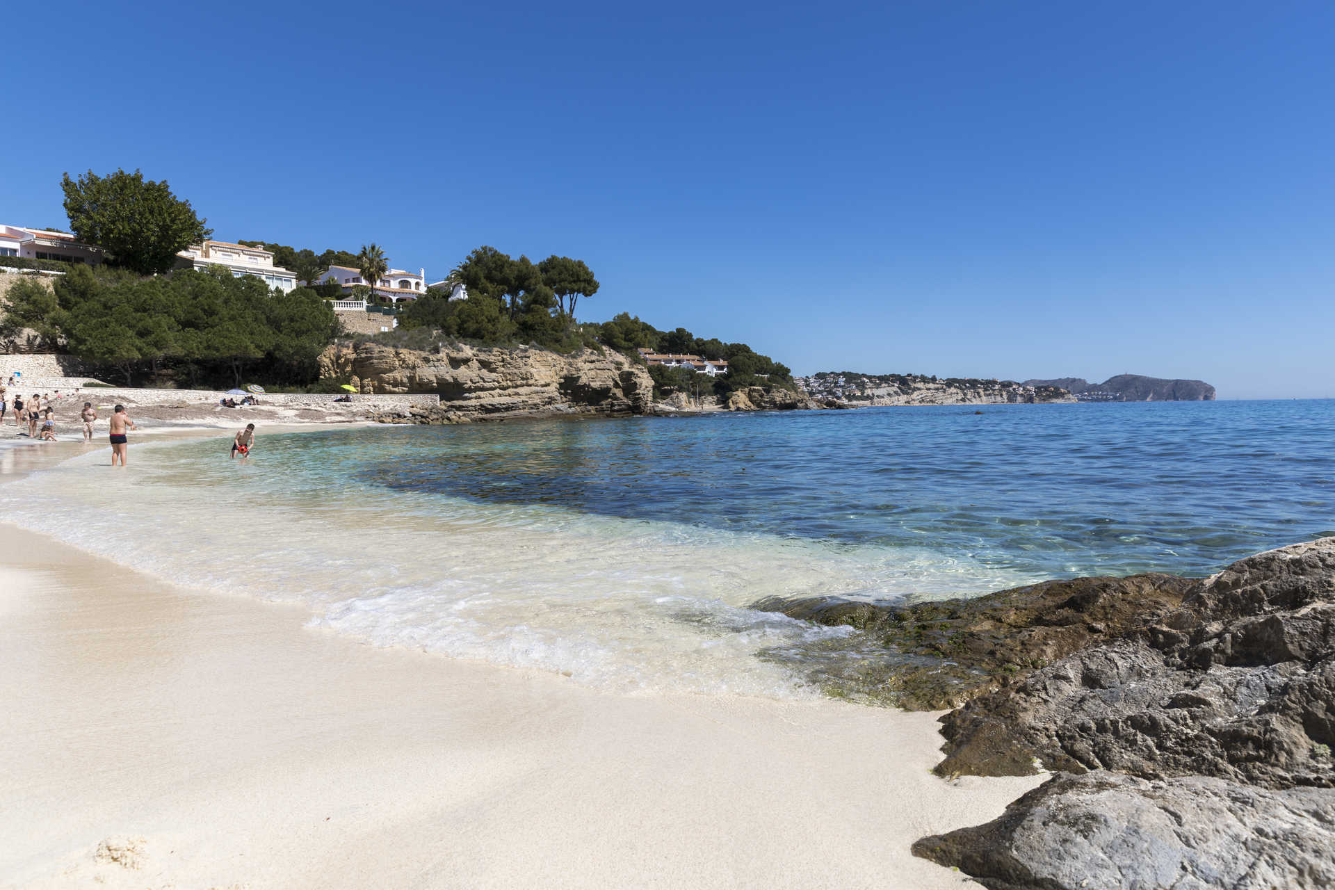 White sandy beach with turquoise waters at La Fustera, featuring rocky outcrops and Mediterranean pines along the coastline under a clear sky.