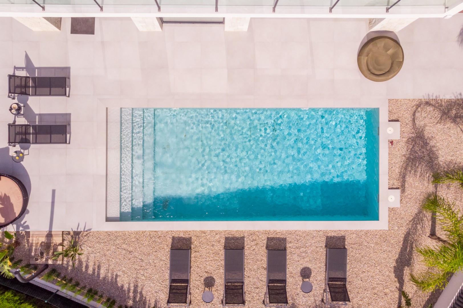 Aerial view of the rectangular pool with steps, surrounded by light tiles and gravel. Loungers and vegetation complete the design.