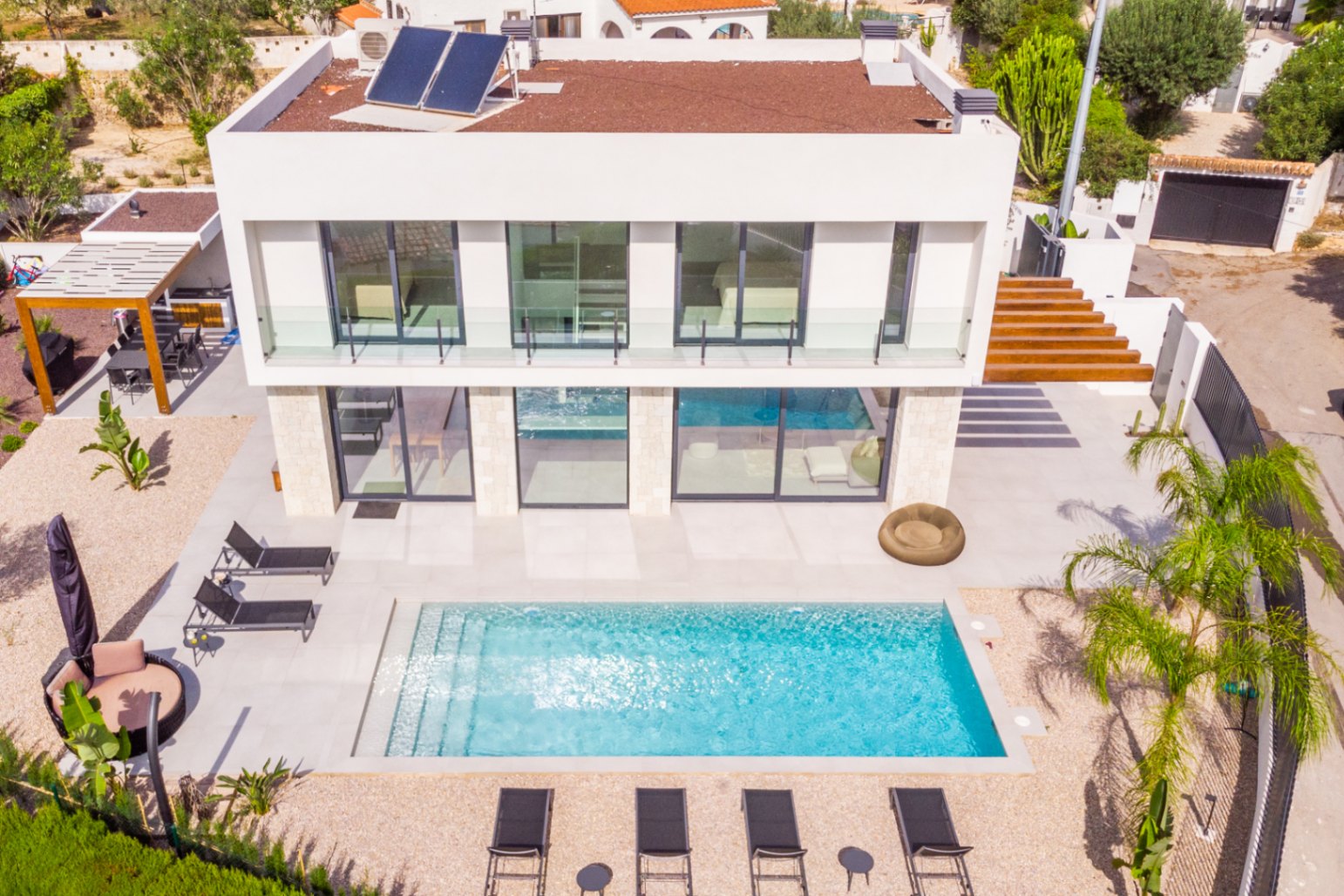 Aerial view of the villa with a rectangular pool, surrounded by sun loungers and light gravel. Modern design with solar panels.