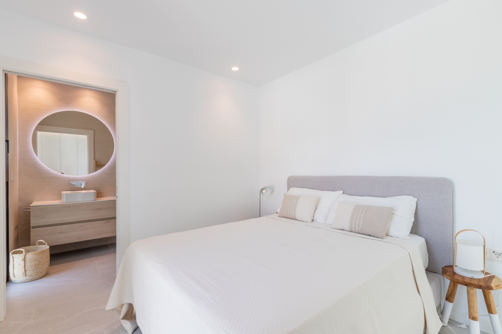 Bright bedroom with ensuite bathroom. Double bed with grey headboard and white linens. Bathroom features a circular illuminated mirror.