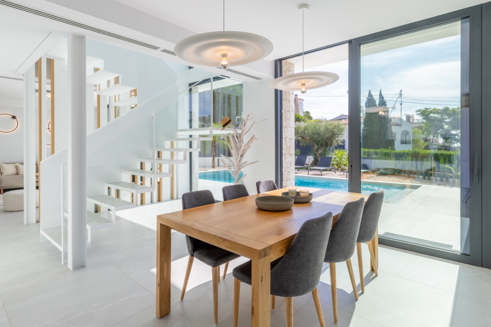 Bright dining room with wood table overlooking the pool and a white floating staircase.