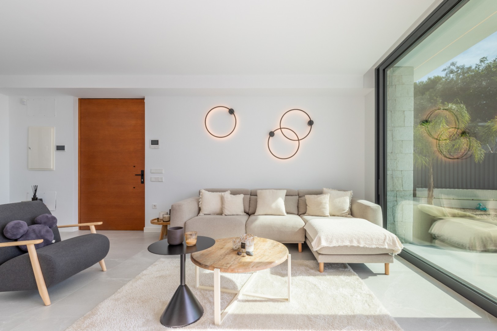 Bright living room with beige sofa, shag rug, and circular lamps. Features a wood door and large window with outdoor views.