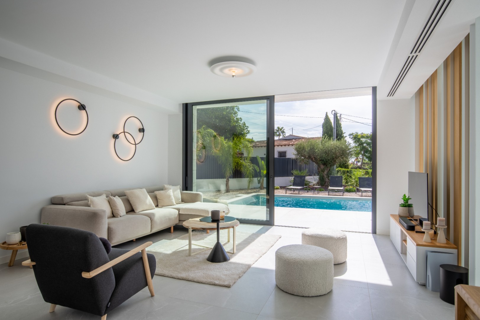 Bright living room with direct pool access, featuring circular wall sconces and modern furnishings.
