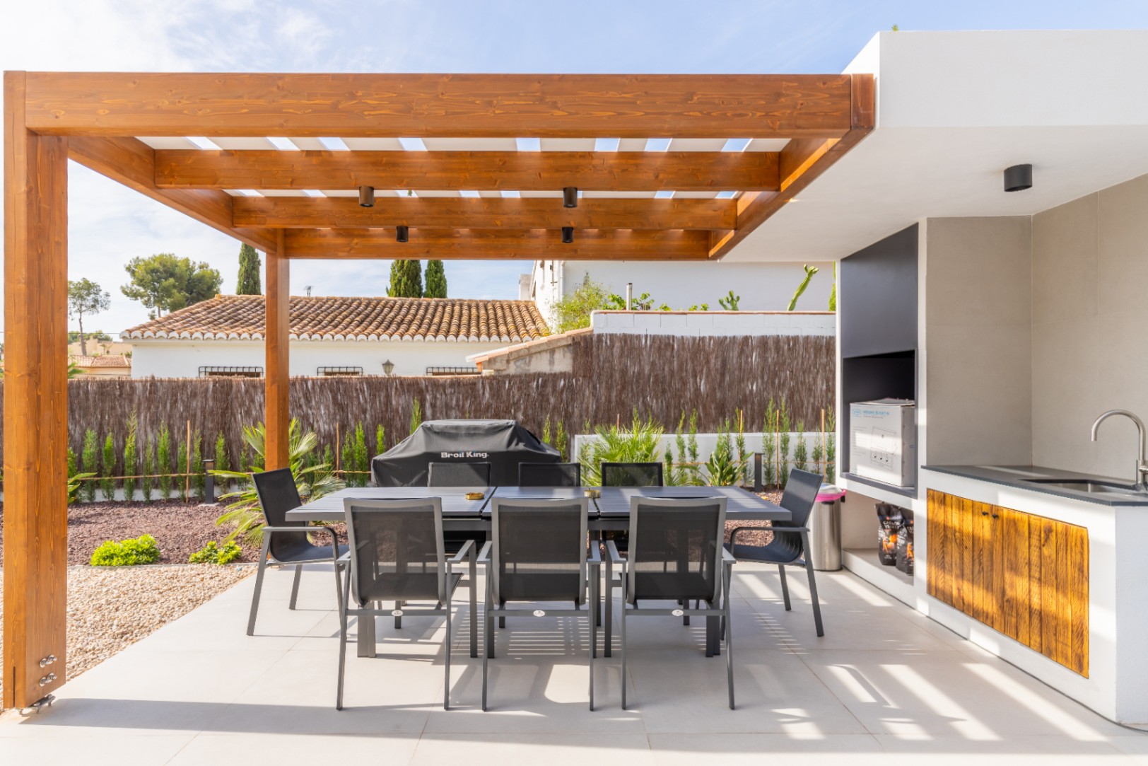 Detail of the outdoor dining area with a wooden pergola and an outdoor kitchen featuring a sink and barbecue.