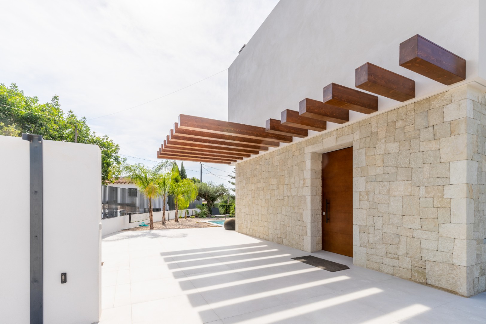 Main entrance featuring a stone wall and a wooden pergola casting shadows on the ground.
