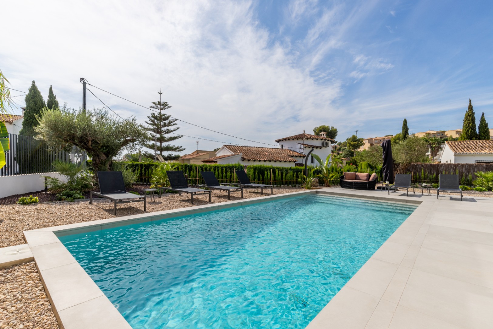 View of the private pool with light stone border, surrounded by black sun loungers and Mediterranean gardens.