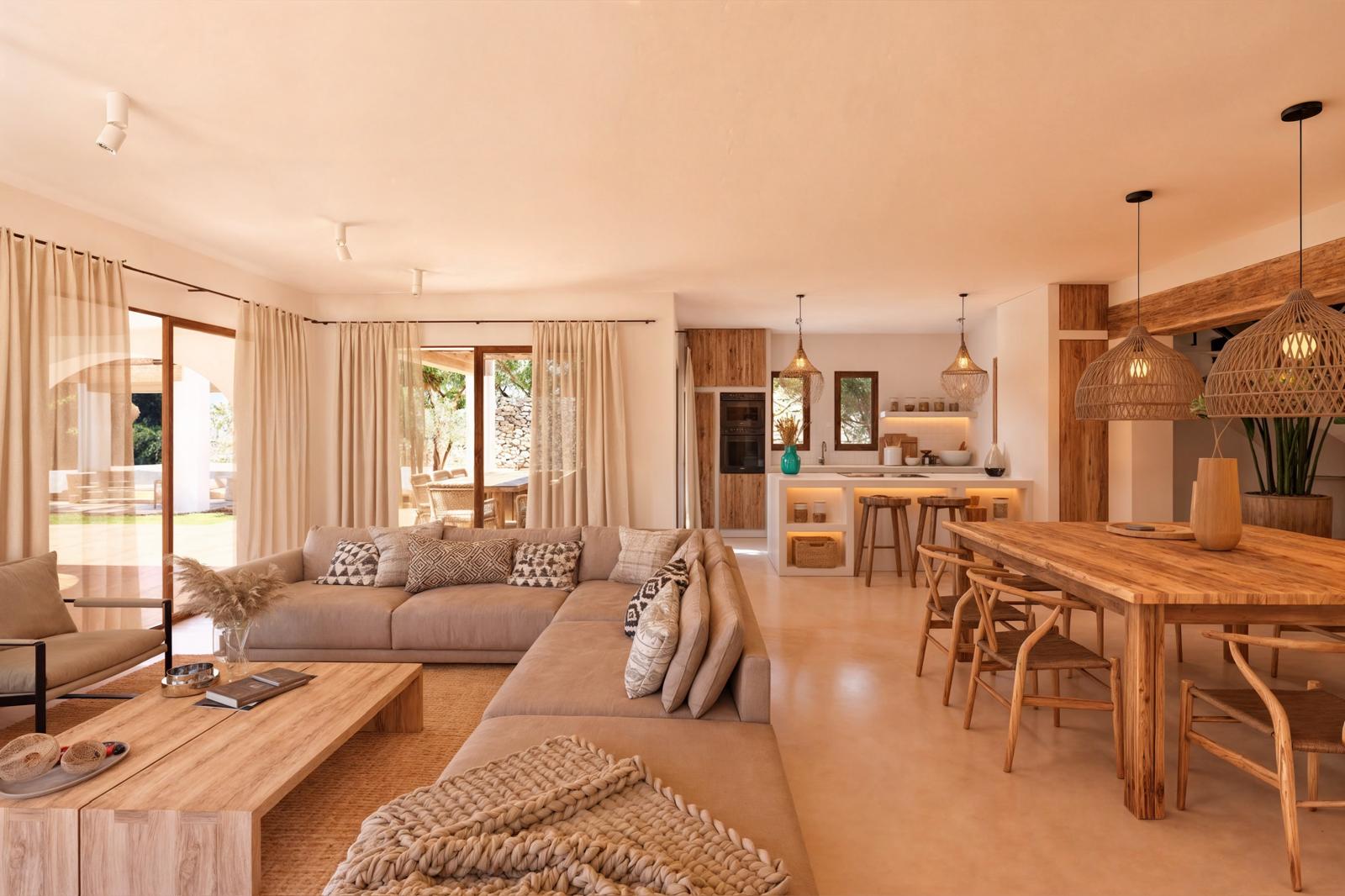 Open-concept living area featuring natural wood furniture, neutral upholstery, and an integrated kitchen with a white island. Wicker pendant lights and large glass doors define the space.