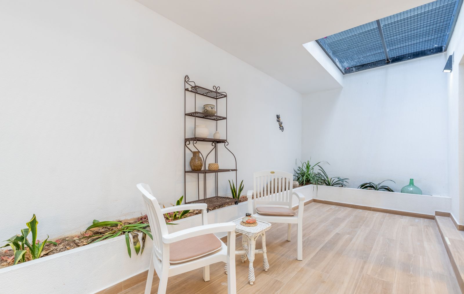 Bright indoor patio featuring a skylight, wood-effect tile flooring, integrated planters, and a seating area with white chairs and a wrought iron shelf.