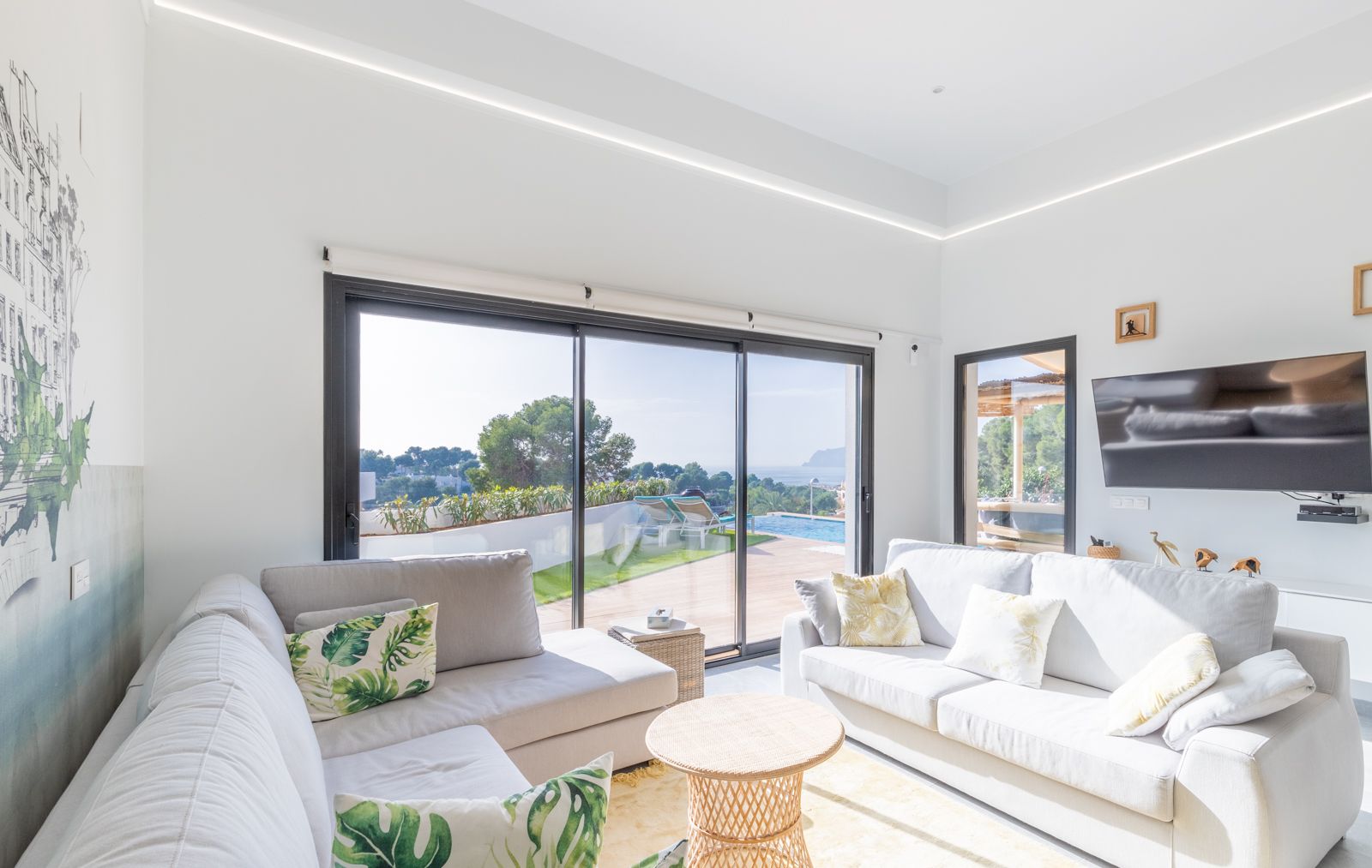 Bright living area featuring grey sofas, integrated LED ceiling lighting, and large sliding doors opening to the terrace with pool and sea views.