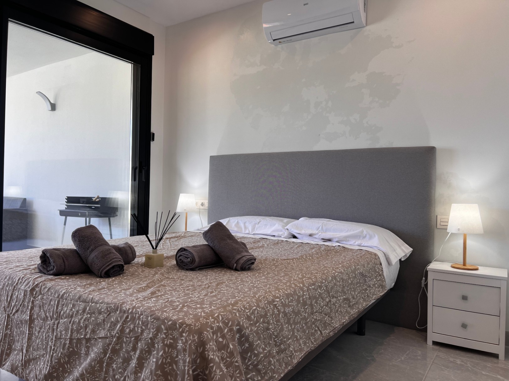 Contemporary bedroom featuring a grey fabric headboard, split-unit air conditioning, and direct terrace access through large black-framed sliding doors.