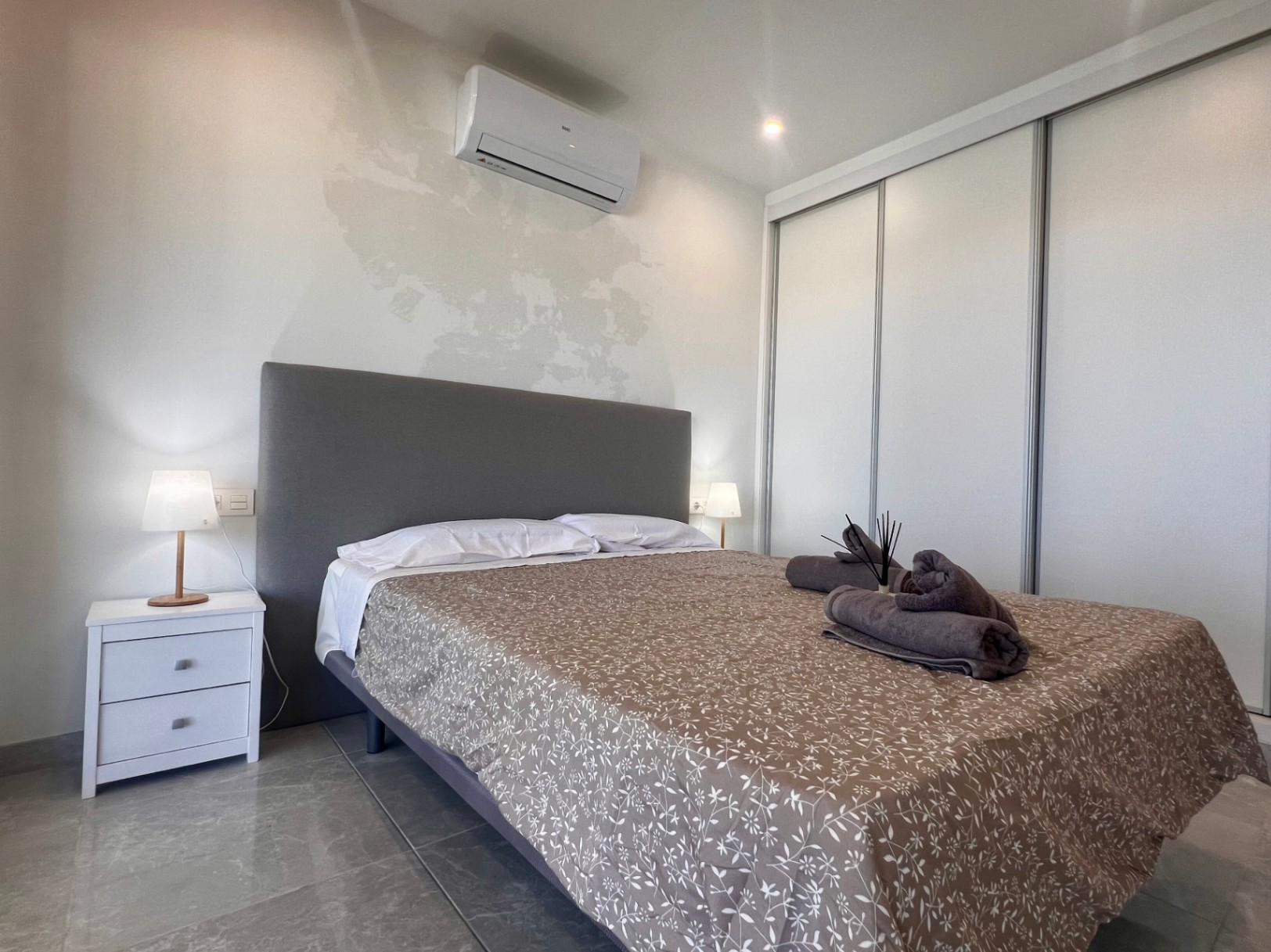 Contemporary bedroom featuring white sliding wardrobes, split air conditioning, and a grey upholstered headboard on polished tile flooring.