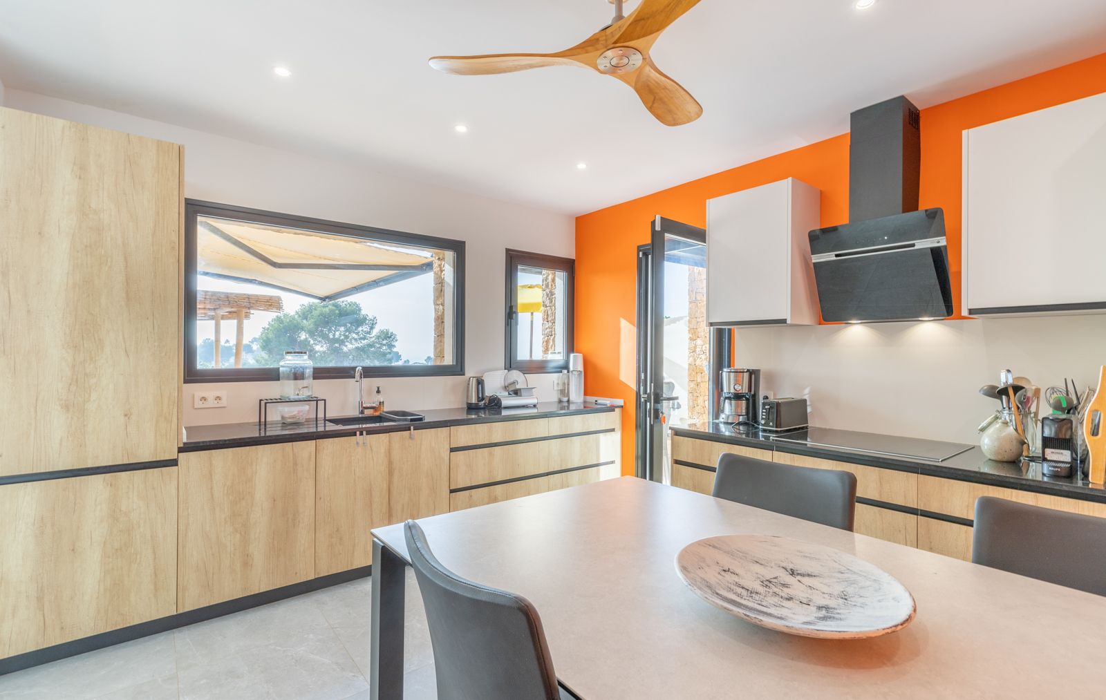 Contemporary kitchen featuring light wood cabinetry, black stone countertops, and a bold orange accent wall. Includes a wooden ceiling fan and windows with terrace views.