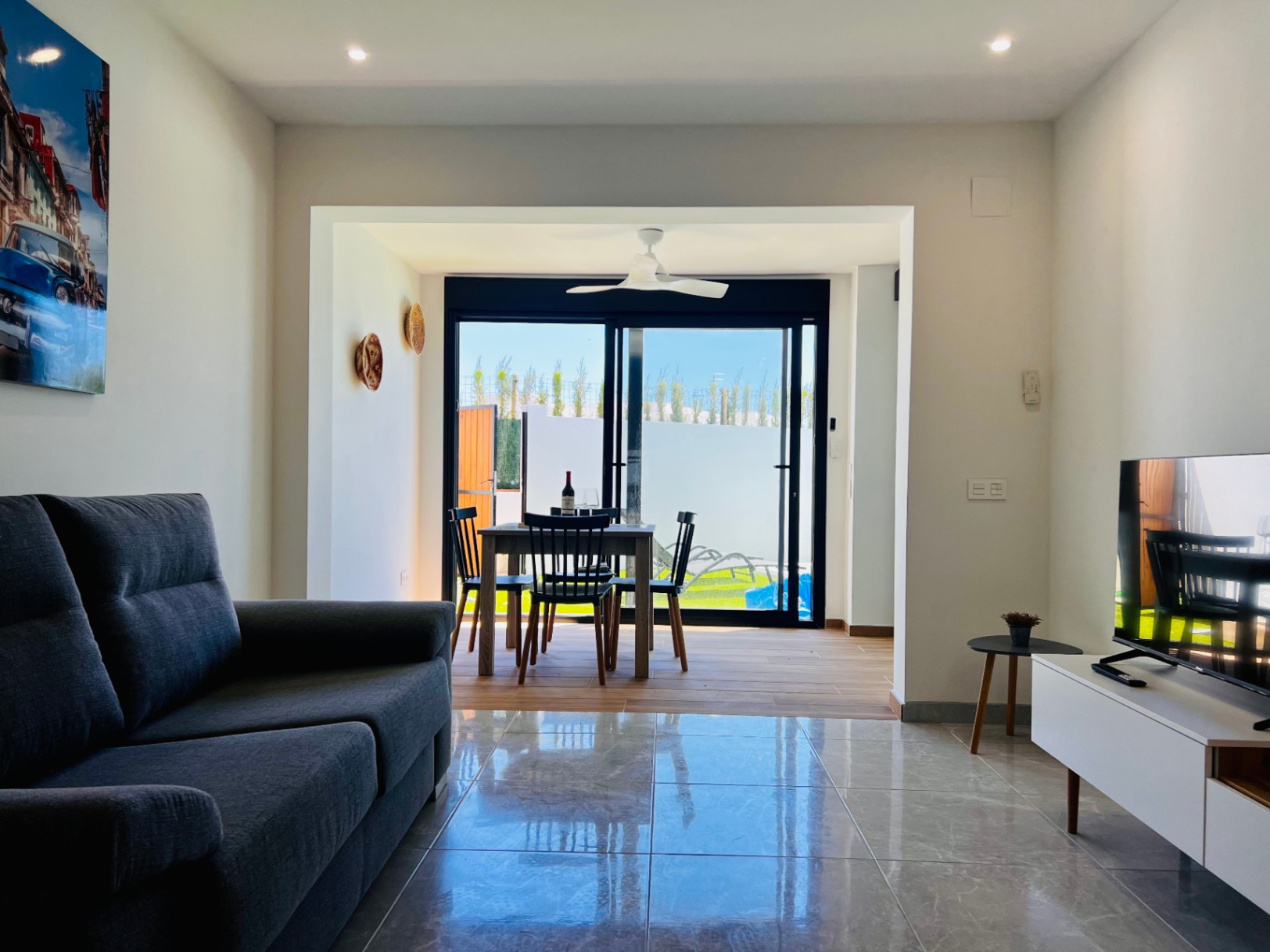 Contemporary living room featuring polished tile flooring, a grey sofa, and an integrated dining area with a ceiling fan and sliding glass doors to the garden.