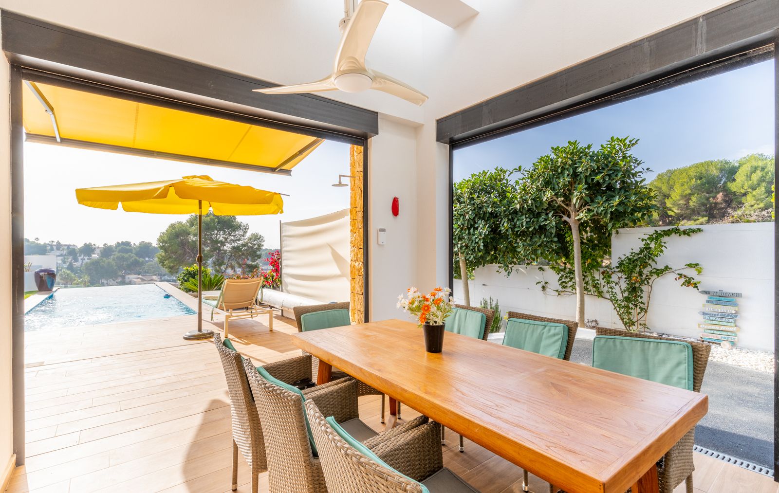 Covered outdoor dining area featuring a wooden table and wicker seating, opening onto the infinity pool deck with integrated ceiling fan and yellow awnings.
