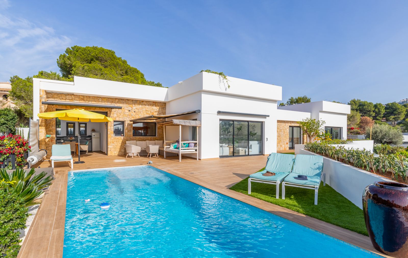 Exterior terrace with a swimming pool, wood-effect porcelain decking, and natural stone walls. Features a summer kitchen, artificial grass solarium, and multiple lounge areas.