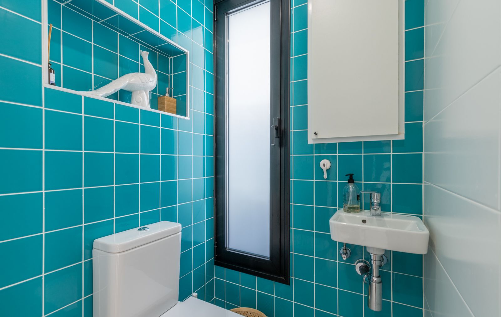 Guest toilet featuring vibrant turquoise wall tiles, a recessed decorative niche, and a compact wall-mounted sink. A slim frosted window provides natural light.