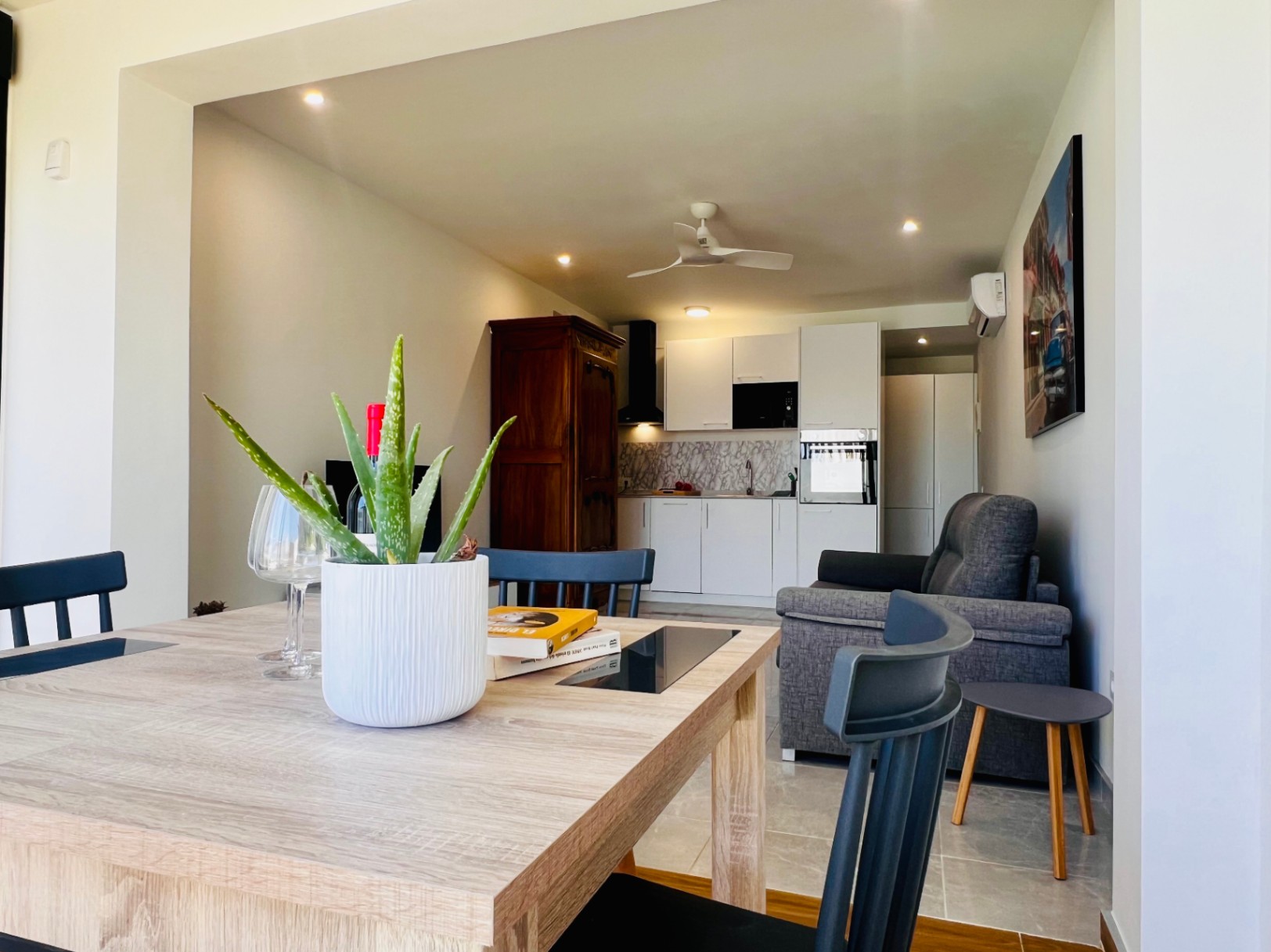 Integrated living-dining area featuring a modern white kitchen, grey sofa, ceiling fan, and a classic wooden armoire providing a warm contrast to the contemporary space.