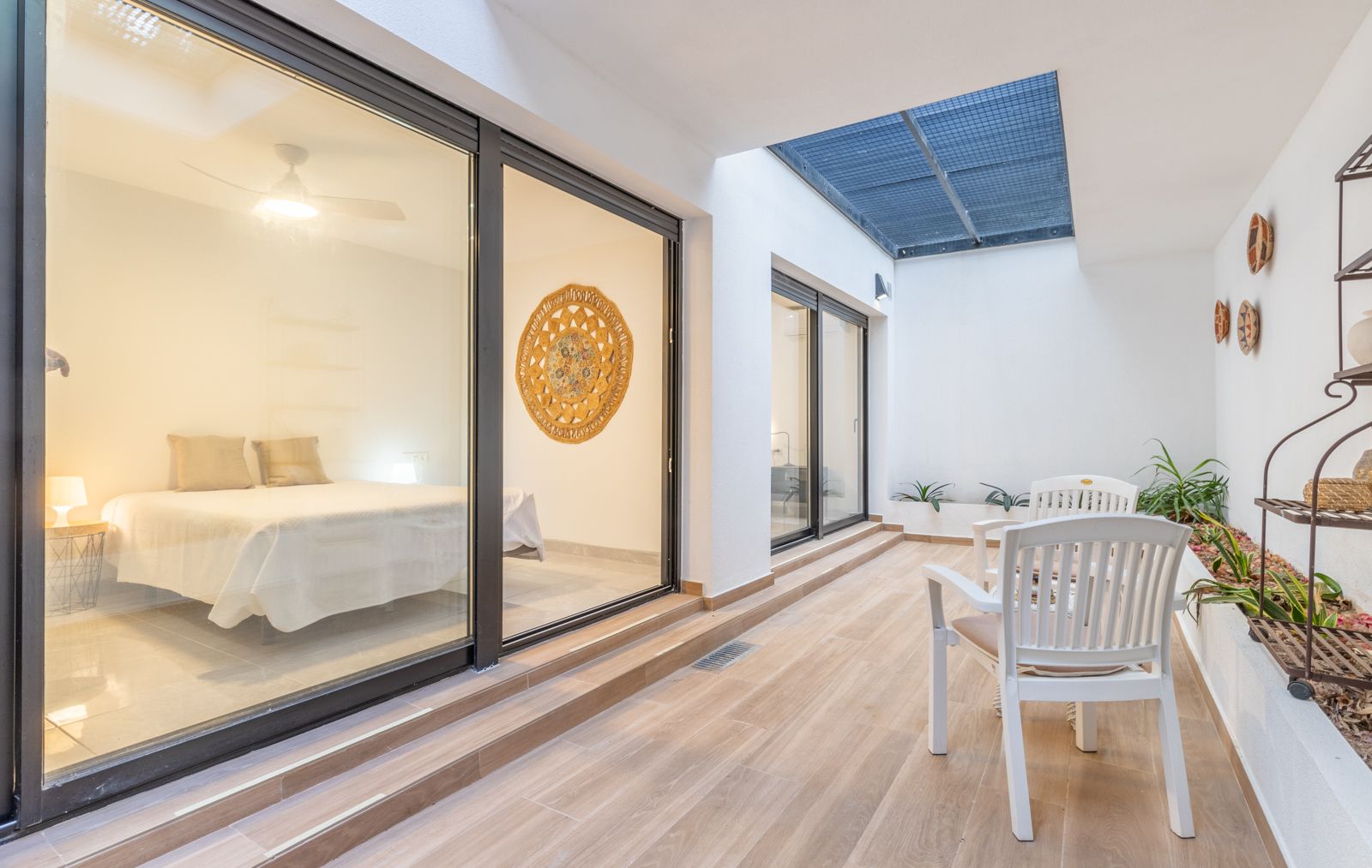 Interior patio featuring wood-effect tiling, a private seating area, and large glass sliders opening into a minimalist bedroom with natural light.