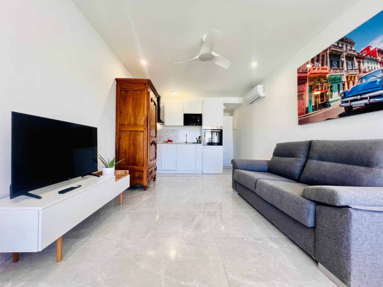 Living area of a guest apartment featuring marble-effect flooring, a modern white kitchenette, grey sofa, and a contrasting antique-style wooden wardrobe.