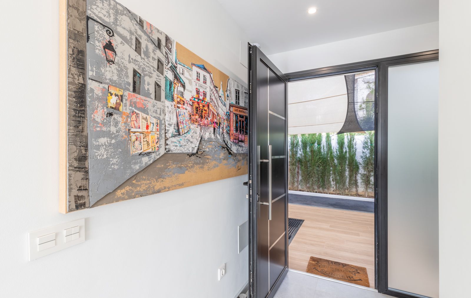 Main entrance featuring a modern anthracite security door, large-scale urban canvas art, and a view towards the exterior wooden decking and privacy hedge.