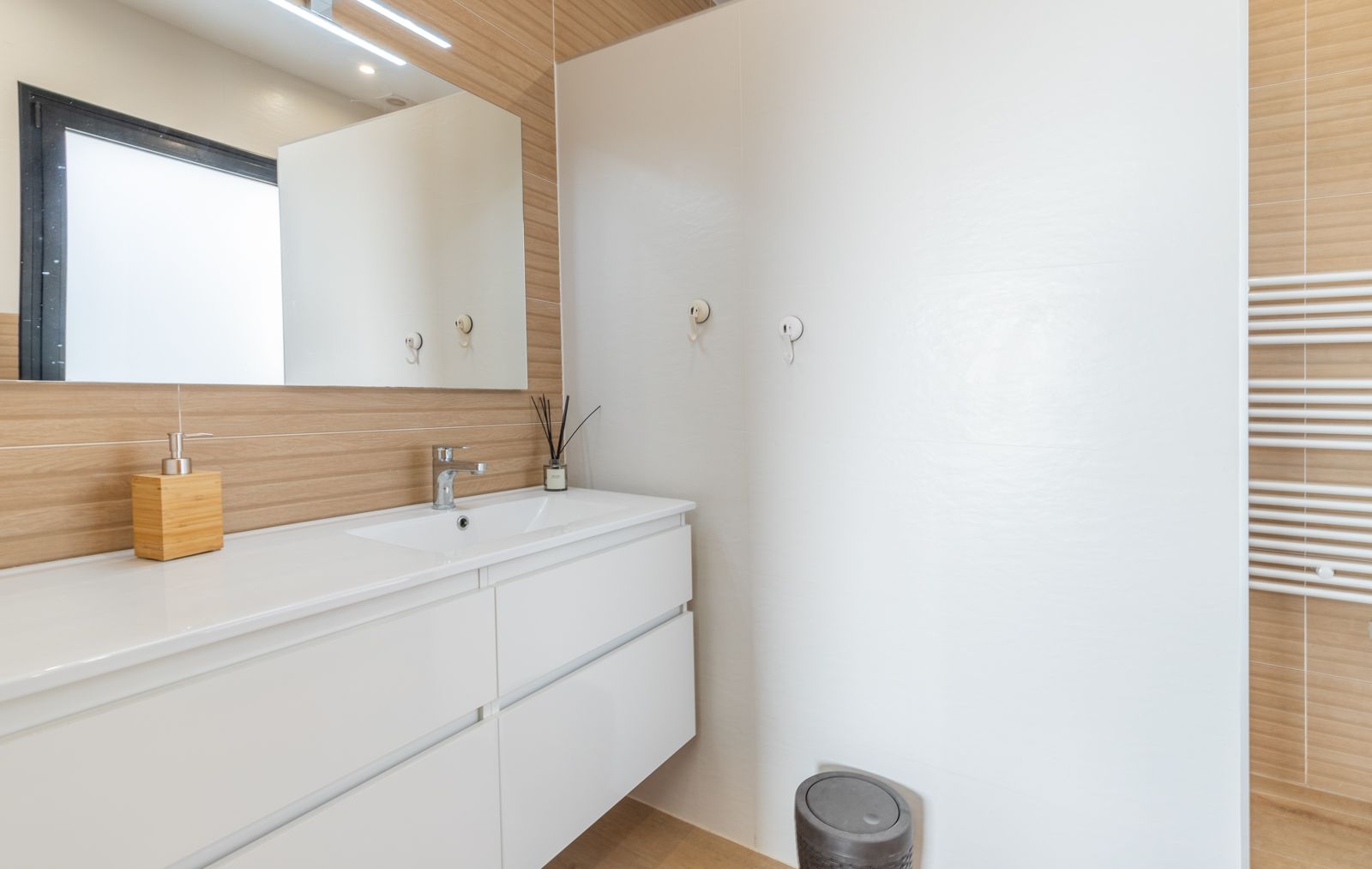Modern bathroom featuring a floating white vanity with integrated sink, wood-effect wall tiling, and a large mirror with overhead LED lighting and a heated towel rail.